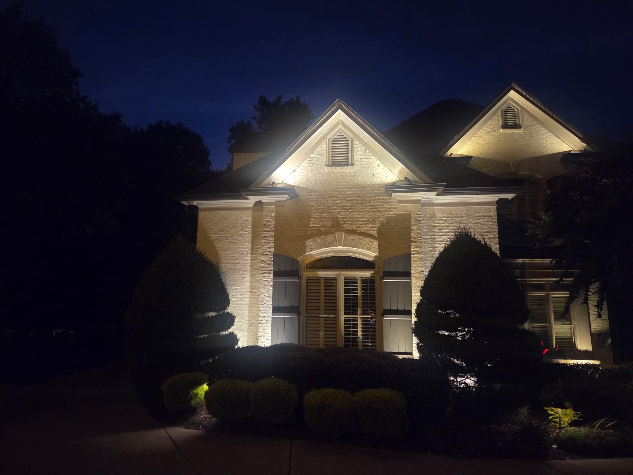 Stone-front home in Dacula, GA 30019 at night with dramatic warm uplighting on the peaked gable, entry columns, and arched doorway, framed by manicured topiary evergreens