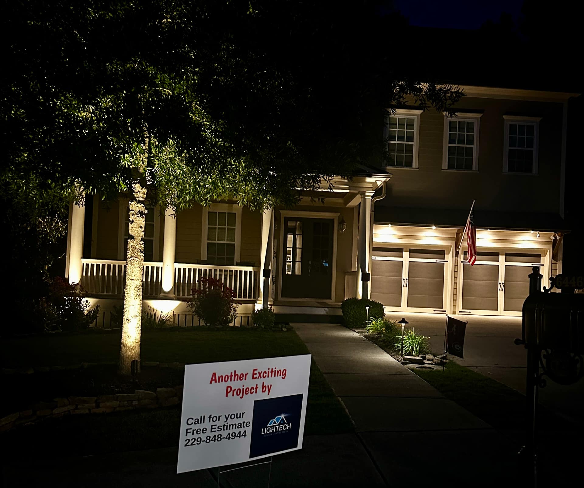 Two-story home at night with warm uplighting and a Lightech project yard sign in the foreground